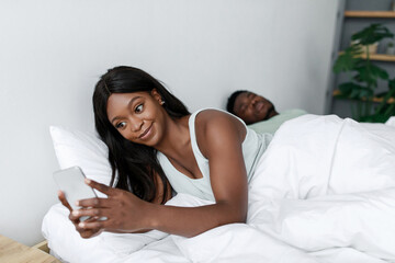 A relaxed woman lies in bed, engaging with her smartphone while her partner sleeps nearby. Sunlight filters through the room, creating a cozy morning vibe.