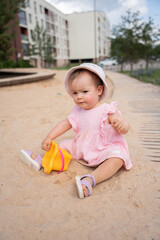 adorable child in pink dress and hat plays happily in sandy urban playground with yellow toy in front of modern apartment buildings and green trees under blue sky, explore, charming, background