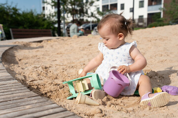 a young child in a white dress sits on a sandy playground, engaged in play with sand toys, under sunlit conditions, surrounded by urban greenery and modern architecture, playful, build, explore