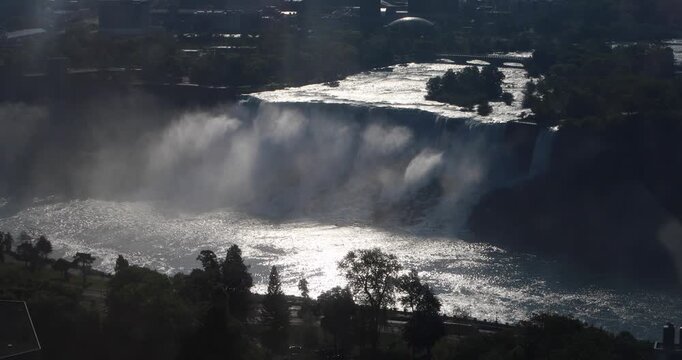 Aerial high angle wide view of the American Falls at Niagara at golden hour. Sunlit water glows silver as thick mist drifts across the river, with the treeline and city in silhouette.