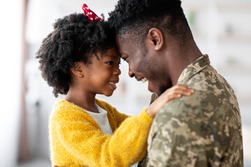 A father in military uniform shares a touching moment with his daughter at home, smiling and embracing each other with love. Their bond shines through in this joyful reunion.