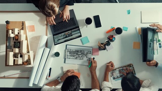 Top view of smart architect engineer writing at blueprint while sitting at meeting table with house model and laptop. Skilled manager working and taking about about building structure. Alimentation.