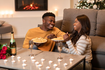 Cheerful black spouses enjoy a romantic dinner with plates of pasta, celebrating the holiday season in their cozy living room, illuminated by candlelight and decorated for Christmas.