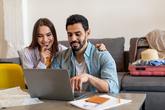 A man and woman are happily planning their trip together at home. They are seated on a couch with a laptop open, surrounded by travel documents and bags.