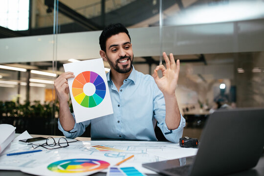 A man smiles and holds up a color wheel during a design meeting. He is reviewing plans on a laptop. The workspace is bright and modern with color samples nearby. - Powered by Adobe
