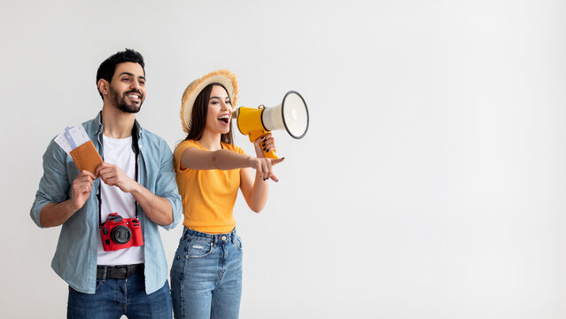 A couple stands cheerfully, showing their travel tickets and a megaphone. The woman points excitedly, while the man smiles happily. They are dressed casually and ready for an adventure.