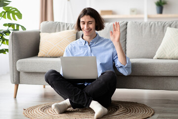 In a bright living room, a young man sits cross-legged on the floor, smiling as he waves during a video call. Soft natural light fills the space, creating a cozy atmosphere. © Prostock-studio