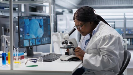 African american woman analyzing molecular samples in a bio lab, using professional microscope. Healthcare and biochemistry experiments represent scientific method and discovery.