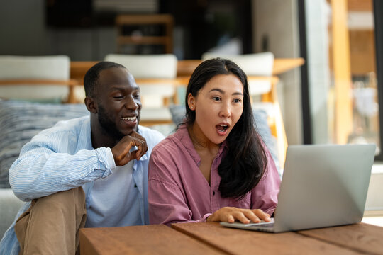 Young happy multiethnic couple having fun while using laptop, digital device at home - Powered by Adobe