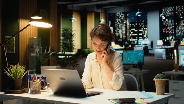 Female assistant engaging in a phone call after hours at the office, giving insight on financial data reports and other metrics for accounting. Chatting on a smartphone connection. Camera A.