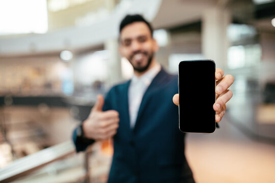 A confident businessman in a suit holds up a blank smartphone while smiling and giving a thumbs up in a bright office environment during daytime.