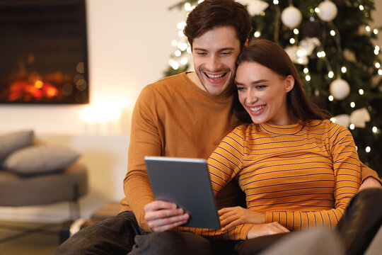 A young couple shares a warm moment while looking at a tablet. They are seated comfortably near a Christmas tree adorned with decorations in a cozy living room setting.