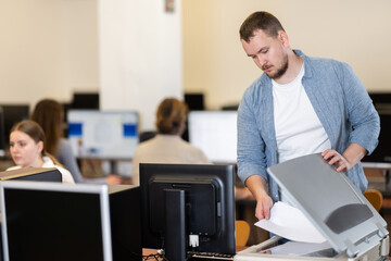 Guy puts paper document into copier, makes copy using office appliances equipment during workday. Office worker stands near copier, figure out how device works