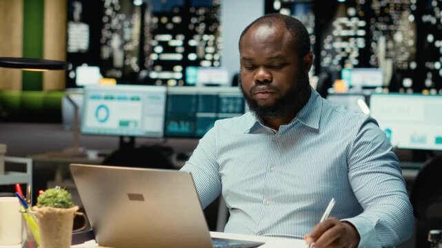 Male finance analyst after hours fact checking and transcribing documents, writing important data on his laptop. Creating a summary and designing strategy to align with company targets. Camera B.
