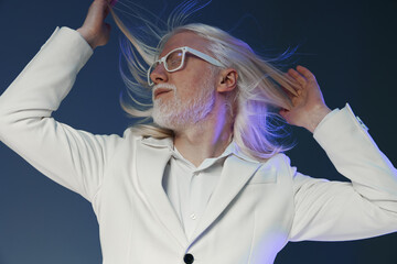 Stylish man with long hair in a white suit, playfully tossing his hair against a gradient...