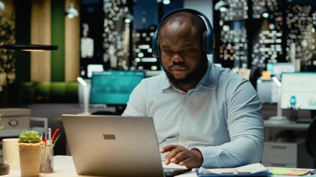 Black stiff man takes break from focus music and stretching in the office, feeling overworked after hours and struggling to stay awake. Sleep deprivation and tension due to overtime. Camera B.