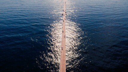 People walk along a wooden pier over calm water reflecting sunlight on a clear day