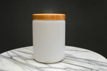 mockup of a white frosted jar, bottle with a wooden lid, sitting on a white marble table with a dark background. Ideal for beauty, cosmetic, food, and packaging design.
