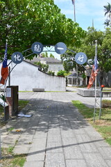 Taiwan - Sep. 20, 2025: Hualong Village entrance with blue circular signs and ROC flags, stone-paved path between grass lawns to white buildings, green trees above, historic settlement, Yilan City.