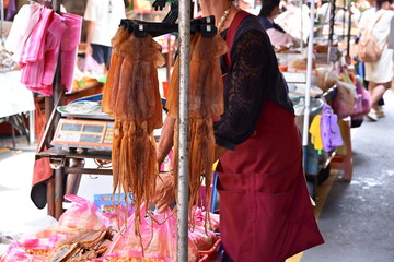 Taiwan - Sep. 20, 2025: Market vendor in black shirt and red apron behind stall with hanging dried brown squid, packaged goods in plastic bags on table, red scale visible, busy market background.