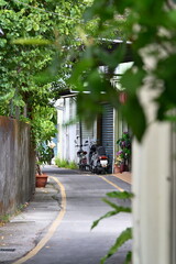 Taiwan - Sep. 20, 2025: Narrow curved alley with yellow line, parked motorcycles, buildings with green plants both sides, wooden fence left, bright peaceful residential scene, Taiwan.
