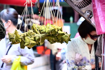Taiwan - Sep. 20, 2025: Hanging zongzi rice dumplings wrapped in green leaves at market stall with two masked women shoppers, traditional Taiwan Dragon Boat Festival food for sale.
