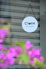 Taiwan - Sep. 20, 2025: White round sign with "Close early tomorrow" and heart on gray shutter, blurred pink-purple bougainvillea in foreground, selective focus composition, Taiwan storefront.
