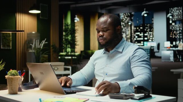 Male finance analyst after hours fact checking and transcribing documents, writing important data on his laptop. Creating a summary and designing strategy to align with company targets. Camera A.