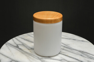 mockup of a white frosted jar, bottle with a wooden lid, sitting on a white marble table with a dark background. Ideal for beauty, cosmetic, food, and packaging design.