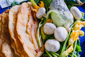 Abundant assortment of fresh Mexican ingredients: quesadillas, huitlacoche, squash blossoms, and mushrooms on a blue background.