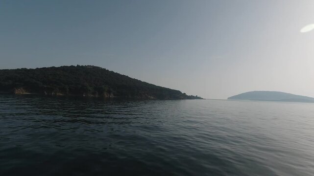 Boat-level tracking shot across shimmering water toward Adalar (Princes&rsquo; Islands). Backlit sun, soft haze, and quiet waves reveal a hillside town and pier, evoking summer calm, distance, and approach.