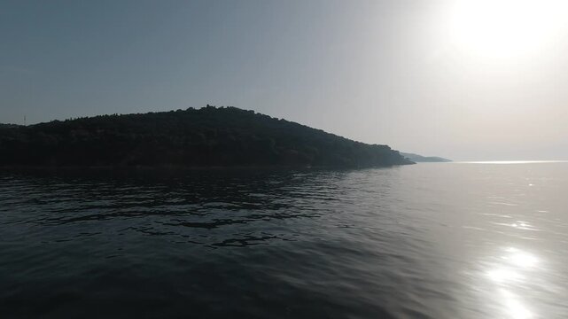 Boat-level tracking shot across shimmering water toward Adalar (Princes&rsquo; Islands). Backlit sun, soft haze, and quiet waves reveal a hillside town and pier, evoking summer calm, distance, and approach.