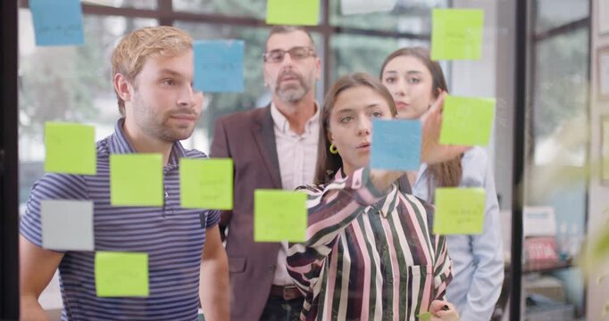 Work colleagues discussing startup project ideas. Team of caucasian business people standing near glass window with sticky notes. Scrum methodology with colorful stickers. 
