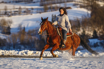 Woman riding brown horse galloping through snowy landscape in winter sunlight