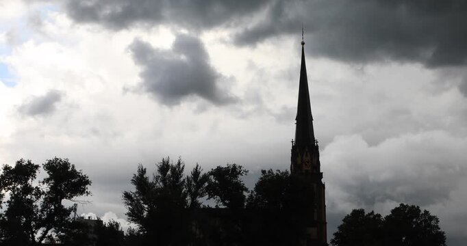 Silhouetted Frankfurt church spire rises beyond dark riverside trees as heavy storm clouds part to reveal a patch of blue. Moody atmosphere, contrast, and a moment of transition after rain.