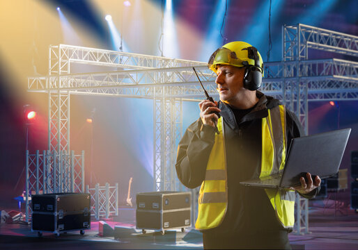 Stage technician with helmet and safety vest coordinates event setup using radio and laptop, supervising lighting and stage construction for live performance.