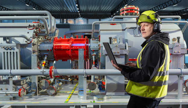Engineer in helmet and safety vest inspects industrial machinery using laptop, monitoring equipment operation and performing technical diagnostics in factory.