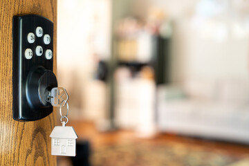 House key with a silver house shaped keychain hanging from a smart lock in a wooden door, open into living room symbolizing welcome or new ownership