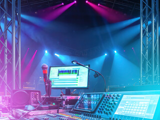 Concert stage control desk with sound mixer, microphone, and computer monitor under colorful stage...