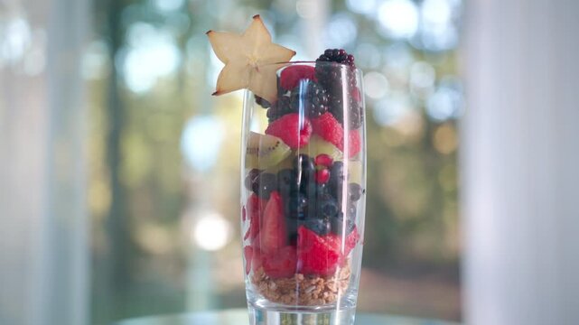 A healthy layered fruit parfait with granola at the bottom, topped with fresh strawberries, blueberries, raspberries, kiwi, blackberries, and a starfruit garnish in a tall glass.