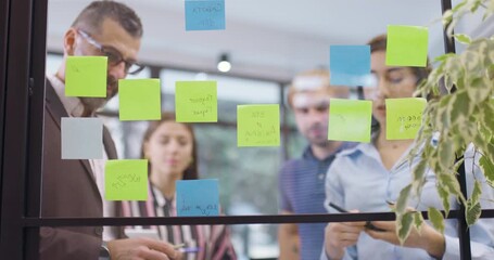 Work colleagues discussing startup project ideas. Team of caucasian business people standing near glass wall with sticky notes. Scrum methodology with colorful stickers. Low angle view focus on window - Powered by Adobe