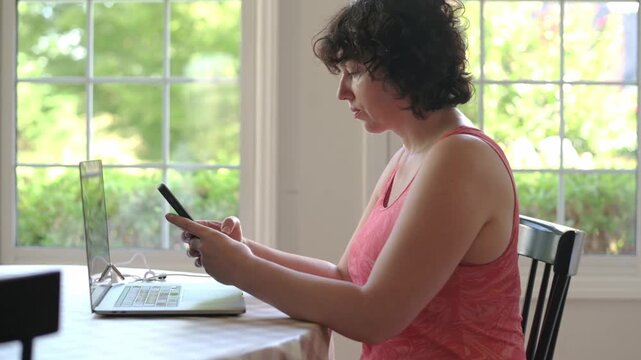 A woman in a pink tank top is sitting at a kitchen table, looking at her phone with a concerned expression. A laptop is open in front of her. 