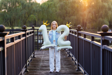 Smiling girl holding birthday balloons in the shape of number twelve on a wooden bridge