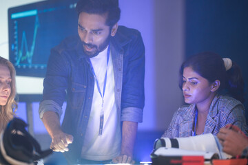 Robotics engineers in an RD lab review a prototype. The Indian man and woman analyze the design on a light table, with AI data graphs on a screen behind them.