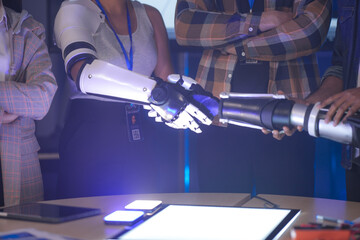 An Indian woman wearing a robotic arm prototype shakes hands with another robotic arm held by a colleague. A diverse tech team smiles, collaborating in a lab.