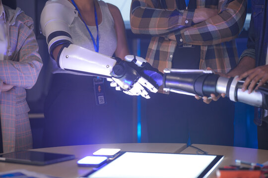 An Indian woman wearing a robotic arm prototype shakes hands with another robotic arm held by a colleague. A diverse tech team smiles, collaborating in a lab. - Powered by Adobe