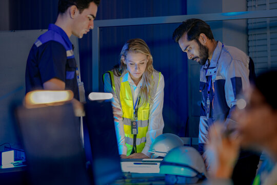 A diverse team of engineers, a Caucasian woman in a safety vest with Indian and Caucasian male colleagues, collaborates late at night. They are focused, reviewing plans in a factory control room.