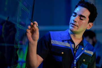 A young, happy Caucasian male engineer in a uniform smiles while using a stylus on a screen. He is working in a high tech control room or factory at night with his team.