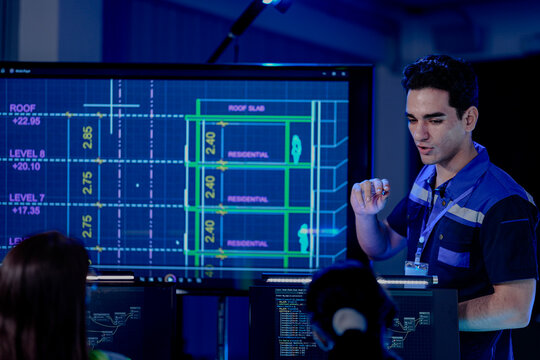 A construction engineer uses a tablet to review a BIM (Building Information Modeling) file. A CAD blueprint of a residential smart building is on a screen in the control center.