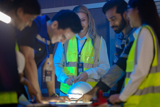 n Indian engineer uses a radio for incident response in a smart factory. His female colleague in a safety vest checks AI system data on a glowing panel in the command center.
